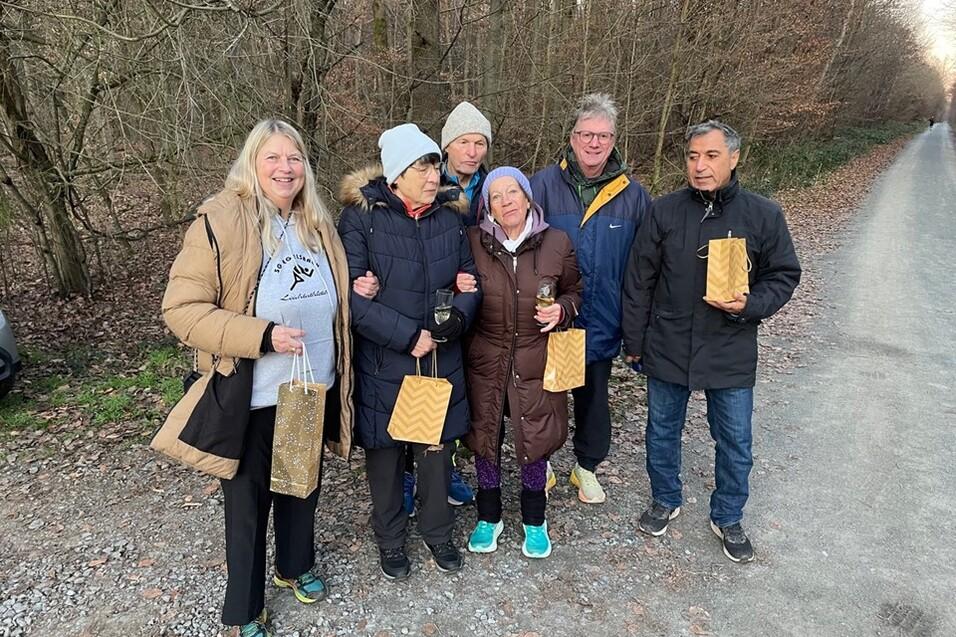 Sechs Personen stehen lächelnd in einer Gruppe auf einem Waldweg, alle halten Geschenktüten in der Hand.
