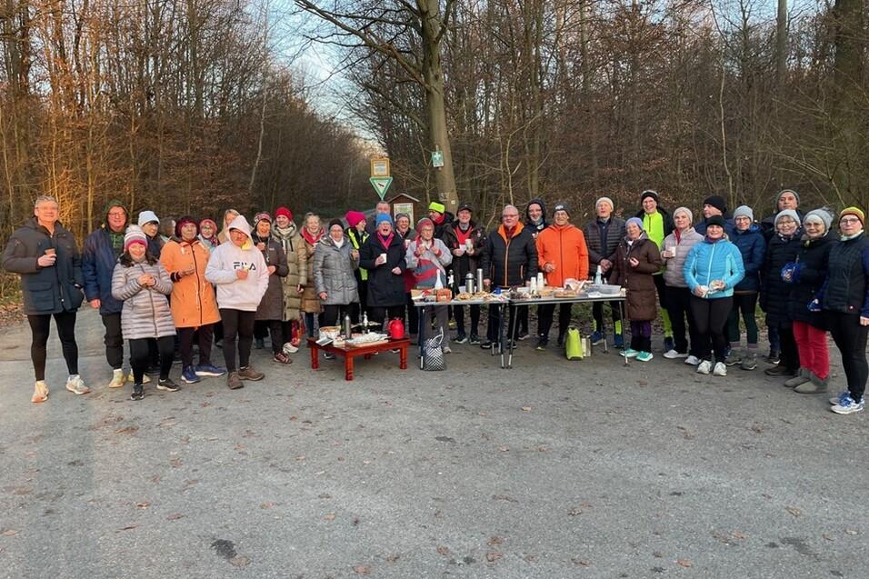 Gruppe von Läufern in Winterkleidung versammelt sich an einem Waldweg, neben einem Tisch mit Getränken und Snacks.