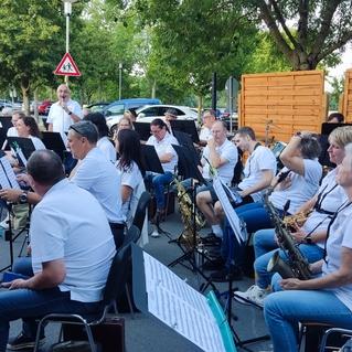 Blasorchester mit Musikern in weißen T-Shirts spielt auf einer Freiluftbühne, umgeben von Zuschauern und Bäumen.