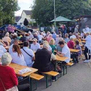 Große Gruppe von Menschen sitzt an langen Bänken und Tischen im Freien, während eine Veranstaltung stattfindet.