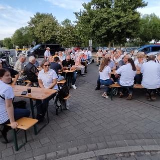 Gruppenszene mit mehreren Personen in weißen T-Shirts, die an Holztischen im Freien sitzen und sich unterhalten.
