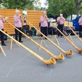 Sechs Musiker spielen Alphörner im Freien, umgeben von Bäumen und einem Transporter.