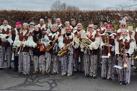 Gruppe von 20 Musikern in gestreiften Kost&uuml;men posiert fr&ouml;hlich vor einer Hecke, Instrumente in der Hand.