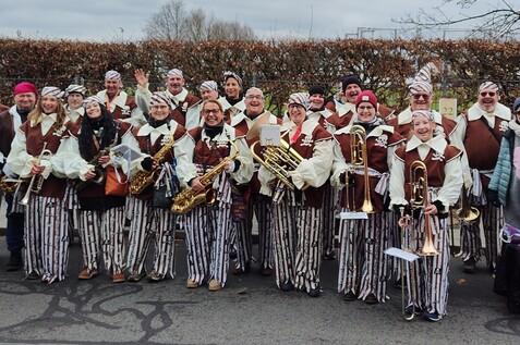 Gruppe von 20 Musikern in gestreiften Kost&uuml;men posiert fr&ouml;hlich vor einer Hecke, Instrumente in der Hand.
