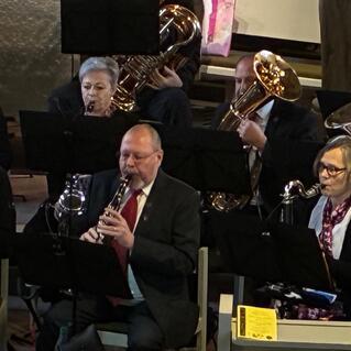 Musikensemble in einem Kirchenraum, Musiker spielen Blasinstrumente, Notenständer sind im Vordergrund sichtbar.