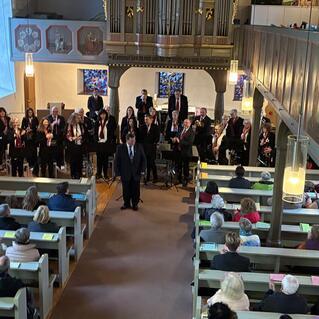 Choraufführung in einer Kirche mit Zuschauern in Bänken und einem Dirigenten auf der Bühne im Vordergrund.