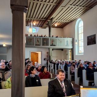 Vollbesetzte Kirche mit Gemeinde im Vordergrund und Empore im Hintergrund, während ein Mann am Klavier sitzt.