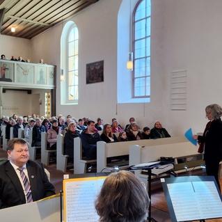 Voller Kirchenraum mit Publikum, einer Rednerin vorne und einer Musikanlage mit Notenblättern im Vordergrund.