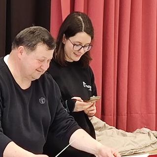 Ein Mann und eine Frau stehen an einem Tisch, während er etwas vorbereitet und sie mit dem Handy fotografiert.