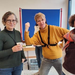 Vier Personen stehen in einem Raum, eine hält einen Becher, während eine andere lächelt und zeigt. Im Hintergrund ist ein blauer Hintergrund sichtbar.