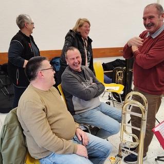 Fünf Personen stehen und sitzen in einem Raum, unterhalten sich und lachen, während Musikinstrumente im Hintergrund sichtbar sind.