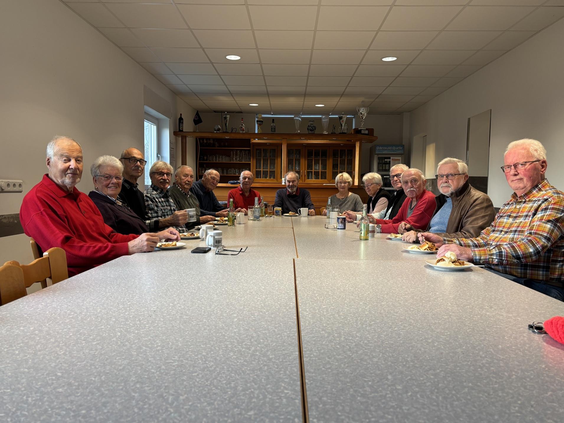 Gruppentreffen von älteren Männern an einem langen Tisch, Snacks und Getränke sind auf dem Tisch verteilt.