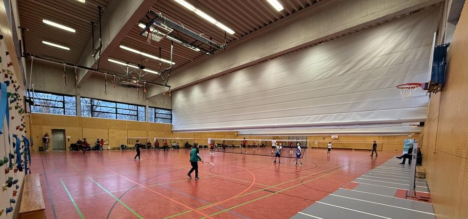 Innenansicht einer Sporthalle mit roten B&ouml;den, Basketballkorb und mehreren Kindern, die Fu&szlig;ball spielen.