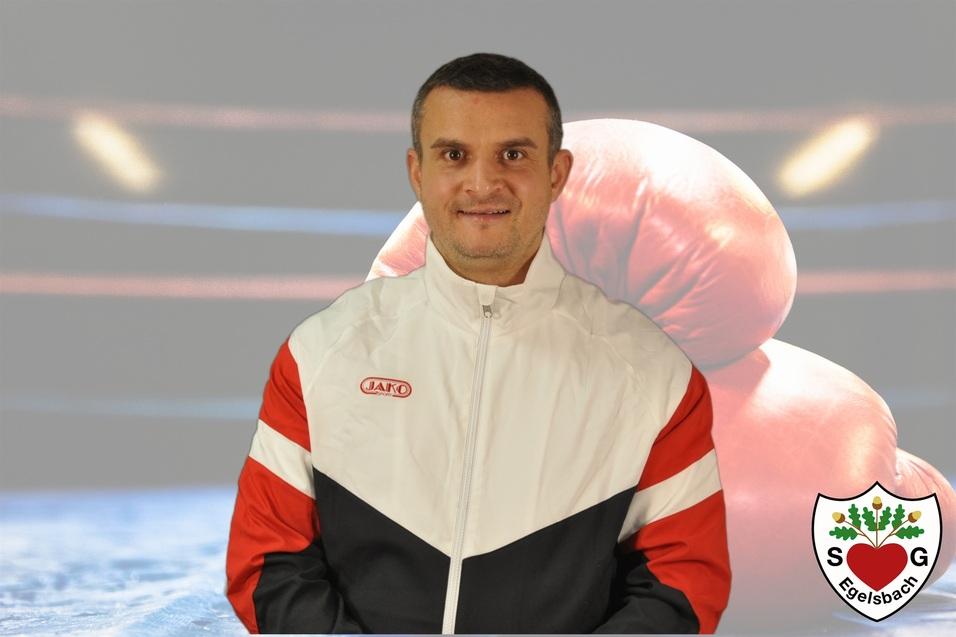 Mann in sportlicher Jacke steht vor einem Boxring mit Boxhandschuhen im Hintergrund. Logo unten rechts.