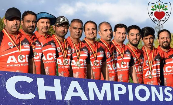 Gruppe von zehn sportlich gekleideten M&auml;nnern mit Medaillen posiert vor einem Banner mit der Aufschrift &bdquo;CHAMPIONS&ldquo;.