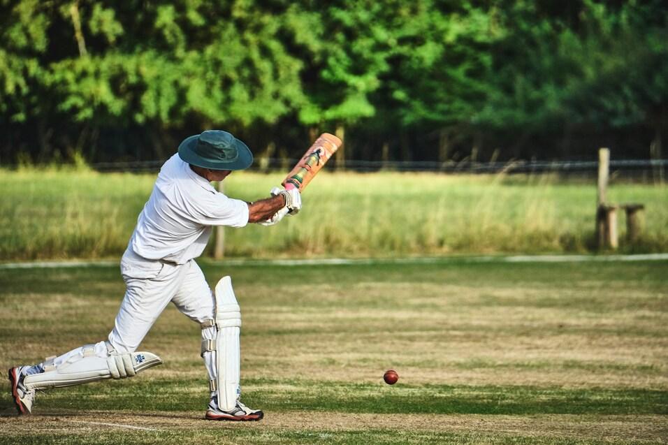 Cricketspieler in wei&szlig;er Kleidung schl&auml;gt mit Schl&auml;ger einen Ball auf einem gr&uuml;nen Spielfeld im Freien.