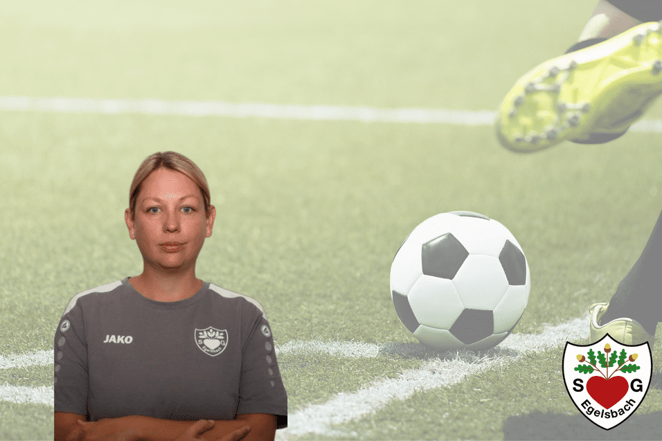 Frau in grauem Sportshirt steht vor einem Fußballfeld, ein Fußball rollt im Hintergrund.