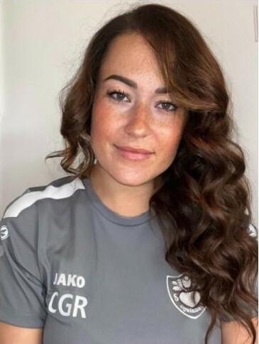 Frau mit lockigem, braunem Haar trägt ein graues Sporttrikot mit Logo und sieht in die Kamera.