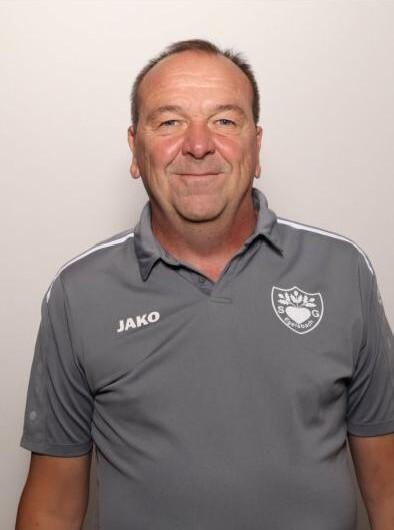 Porträt eines lächelnden Mannes in grauem Poloshirt mit Jako-Logo und Vereinsabzeichen auf heller Hintergrund.