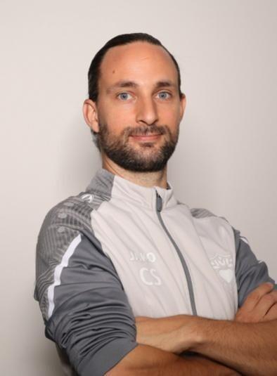 Porträt eines Mannes mit Halbhochfrisur, der in Sportbekleidung mit Logo vor neutralem Hintergrund posiert.