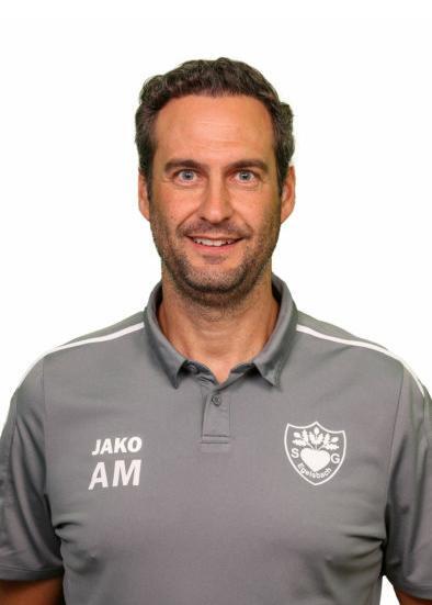 Porträt eines Mannes mit lässigem Lächeln, in grauer Trainerjacke mit Logo und Schriftzug „JAKO AM“.