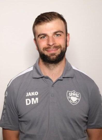 Porträt eines Mannes mit Bart in einem grauen Sportshirt, lächelnd vor einem neutralen Hintergrund.