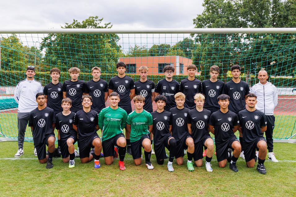 Gruppenfoto eines Fu&szlig;ballteams mit 16 Spielern in schwarzen Trikots, stehend vor einem Tor auf einem Rasenplatz.