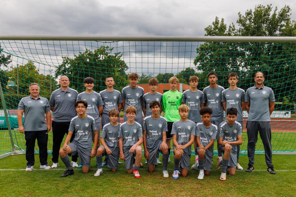 Gruppenshot einer Fu&szlig;ballmannschaft mit Trainern auf dem Platz, alle tragen graue Trikots und stehen vor einem Tor.