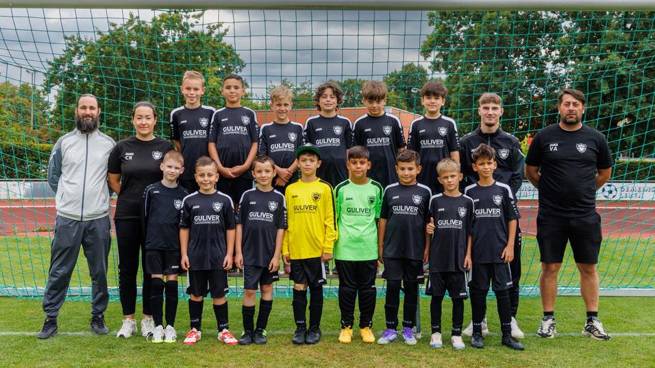 Mannschaftsbild einer Jugendfu&szlig;ballmannschaft in schwarzen Trikots vor einem Tor, umgeben von Trainer und Betreuern.