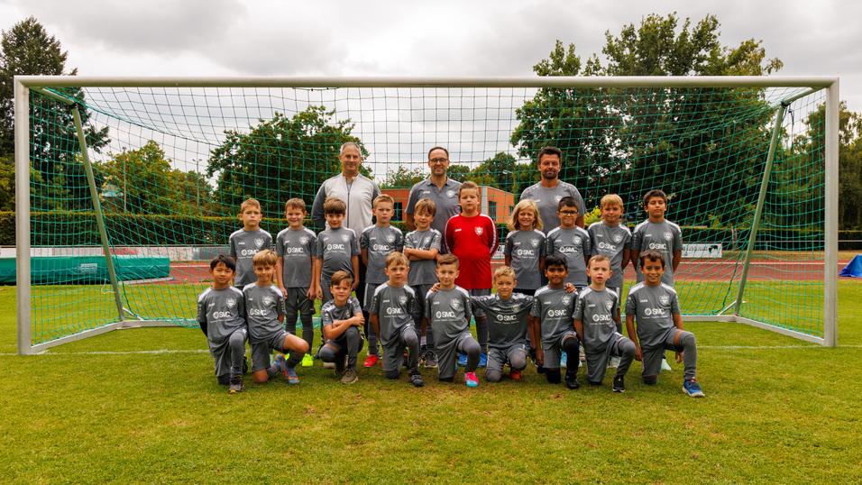 Gruppenfoto einer Fu&szlig;ballmannschaft mit Trainer, 24 Kinder in grauen Trikots vor einem Fu&szlig;balltor auf einer Rasenfl&auml;che.