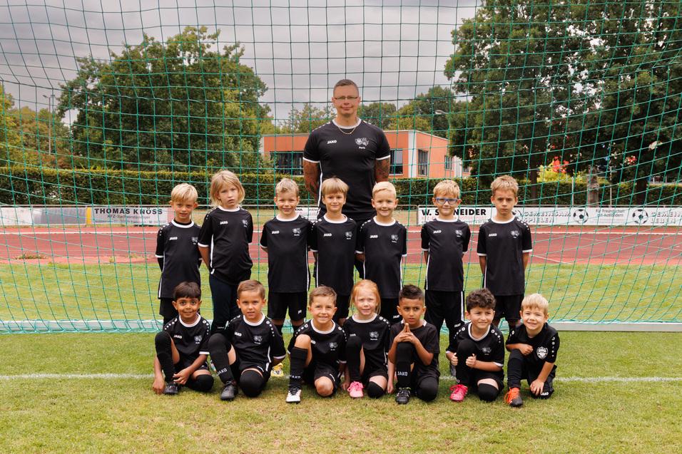 Gruppenfoto einer Jugendfu&szlig;ballmannschaft mit Trainer vor einem Tor, alle tragen schwarze Trikots und stehen auf dem Rasen.