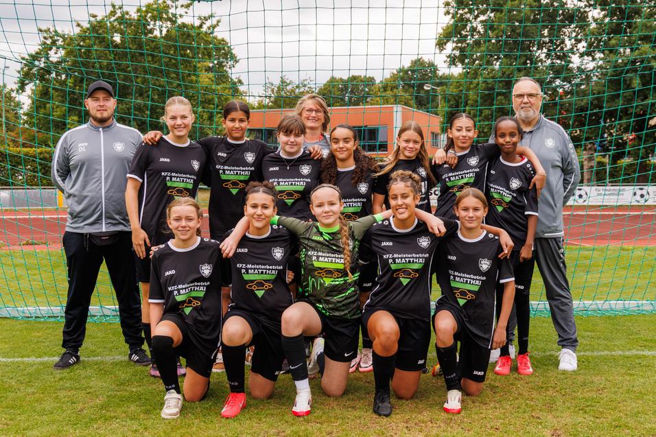 Mannschaftsfoto eines M&auml;dchenteams in schwarzen Trikots, umgeben von Trainern, vor einem Fu&szlig;balltor auf dem Platz.