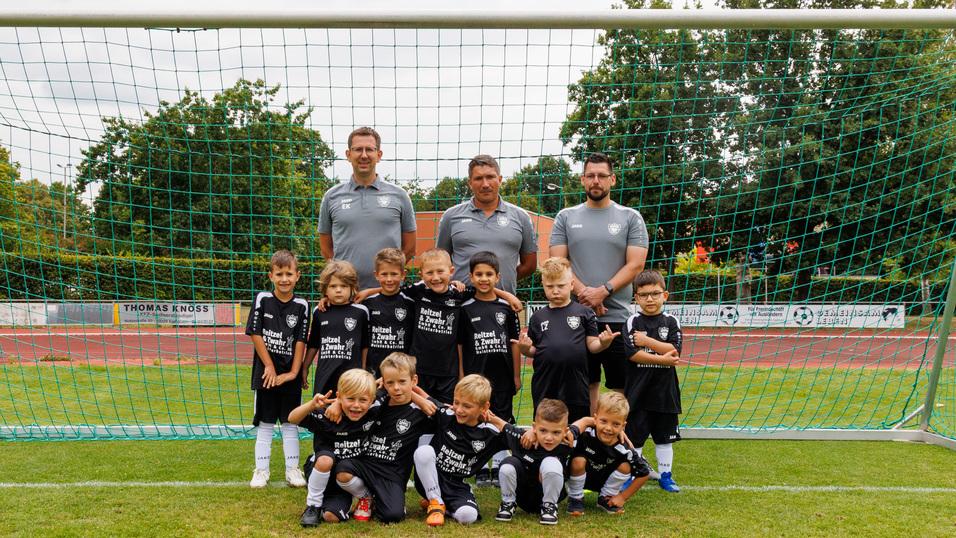 Kinderfu&szlig;ballmannschaft in schwarzen Trikots posiert vor einem Fu&szlig;balltor auf einem Sportplatz. Erwachsene Trainer im Hintergrund.