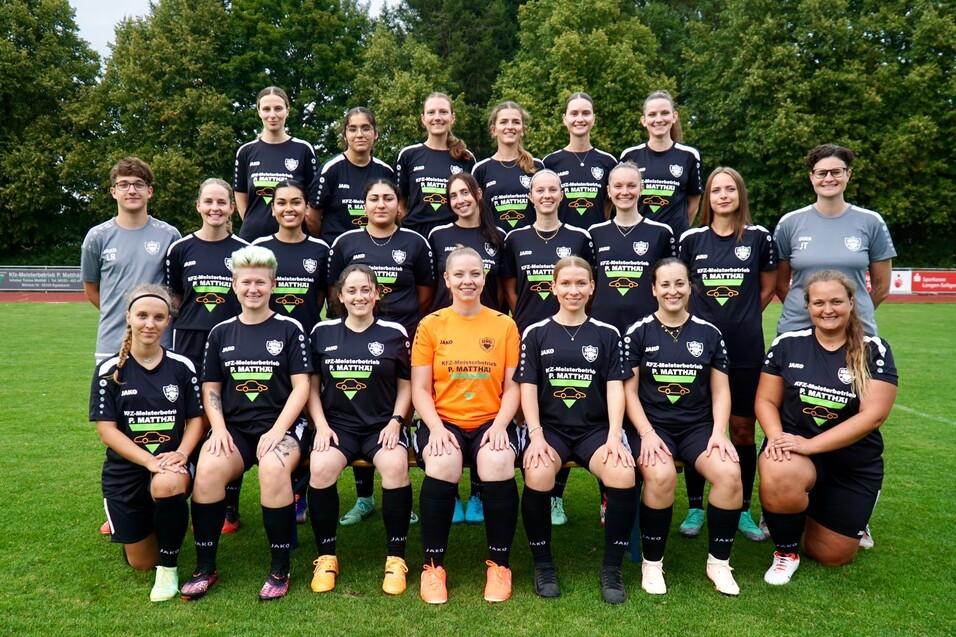 Gruppenfoto einer Frauenfu&szlig;ballmannschaft in schwarzen Trikots auf dem gr&uuml;nen Rasenplatz, 24 Spielerinnen und Trainer.