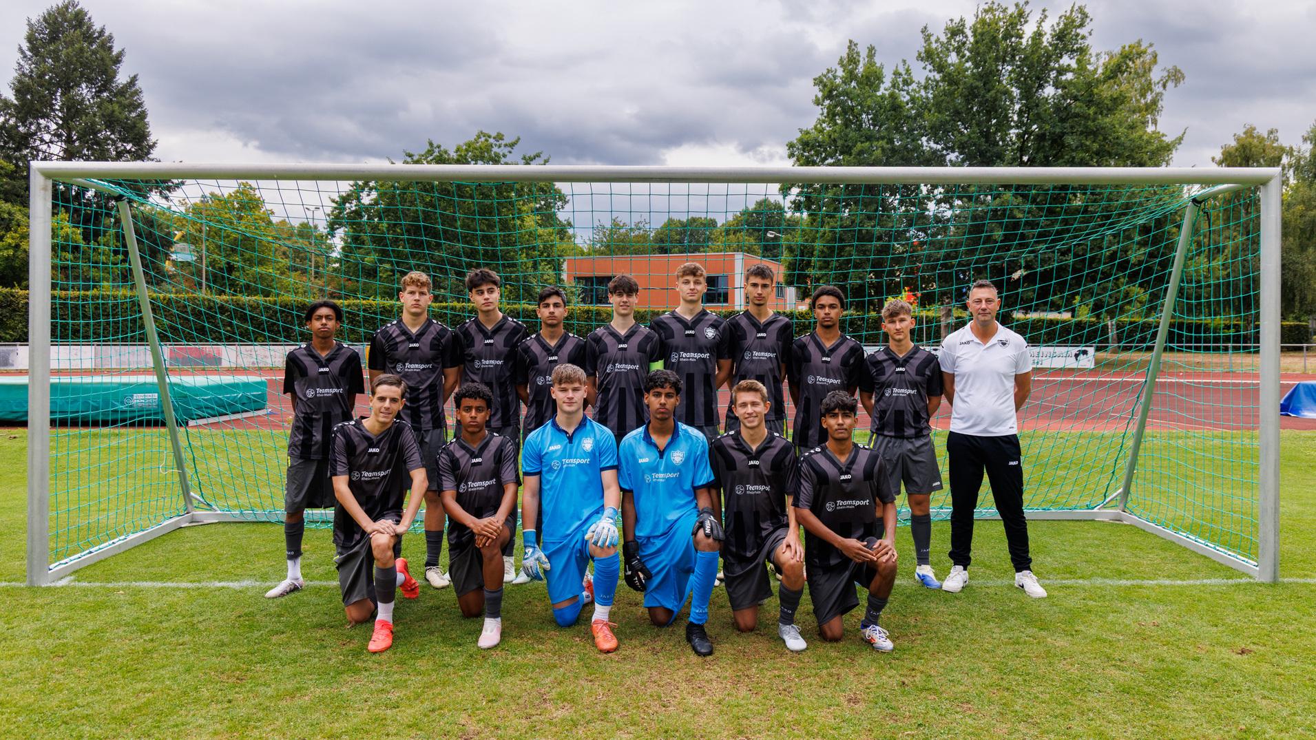 Mannschaftsbild einer Fußballjugend mit Trainer vor einem Tor auf einem Rasenplatz, Jugendliche in dunklen Trikots, ein Torwart in Blau.