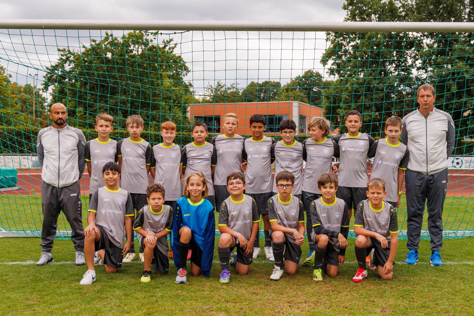 Gruppenfoto einer Jugendfu&szlig;ballmannschaft mit zwei Trainern vor einem Fu&szlig;balltor auf einem Rasenplatz.