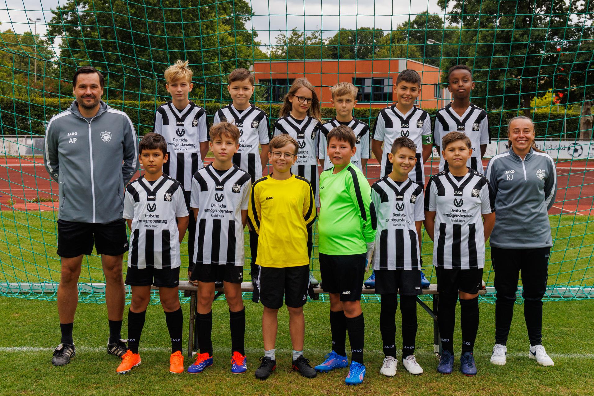Gruppe von zw&ouml;lf Fu&szlig;ballspielern in Schwarz-Wei&szlig;-Trikots und zwei Trainern vor einem Fu&szlig;balltor auf einem Sportplatz.