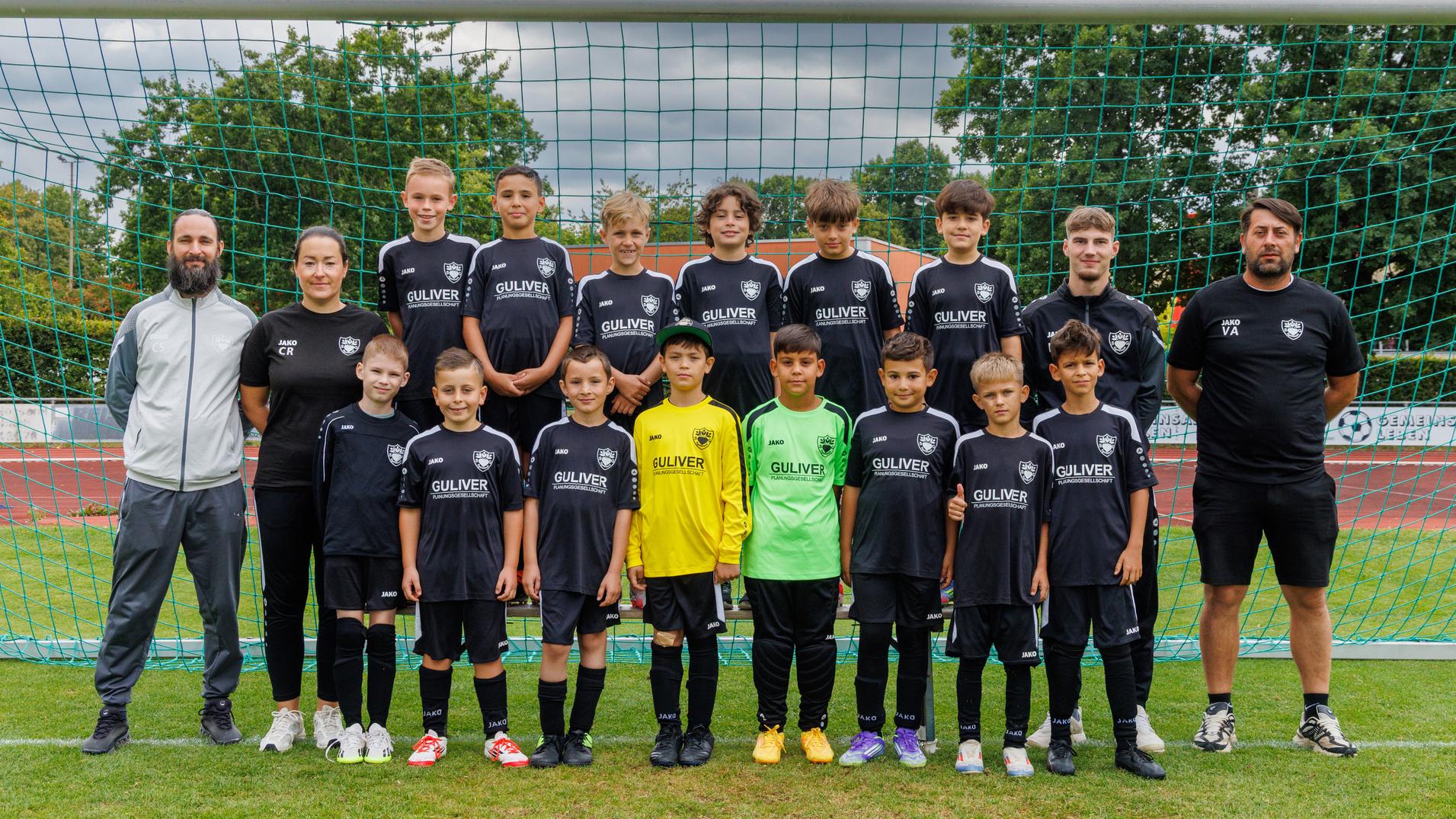 Mannschaftsbild einer Jugendfußballmannschaft in schwarzen Trikots vor einem Tor, umgeben von Trainer und Betreuern.