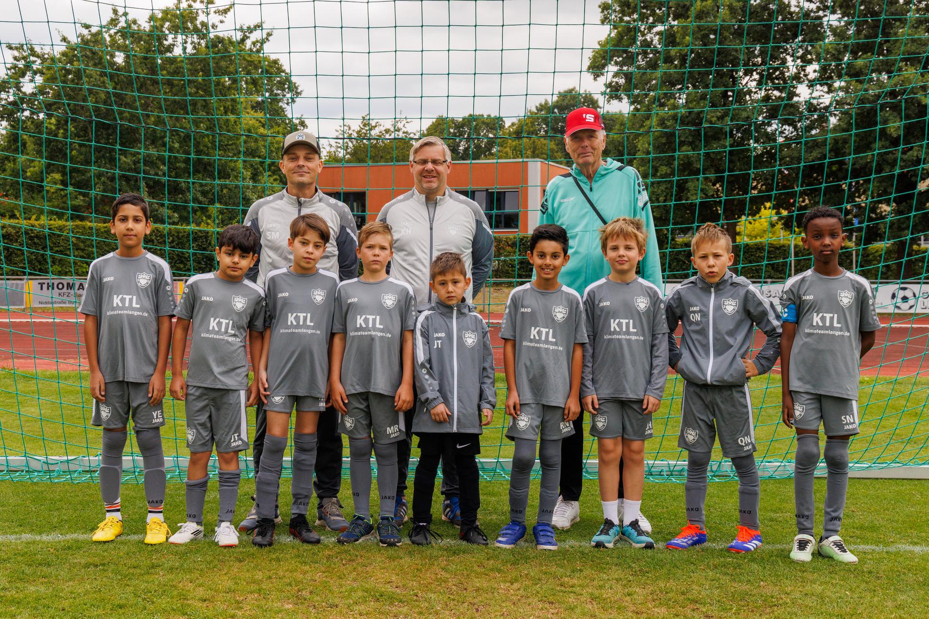 Gruppenfoto einer Jugendfu&szlig;ballmannschaft in grauen Trikots vor einem Fu&szlig;balltor, begleitet von drei Trainern.