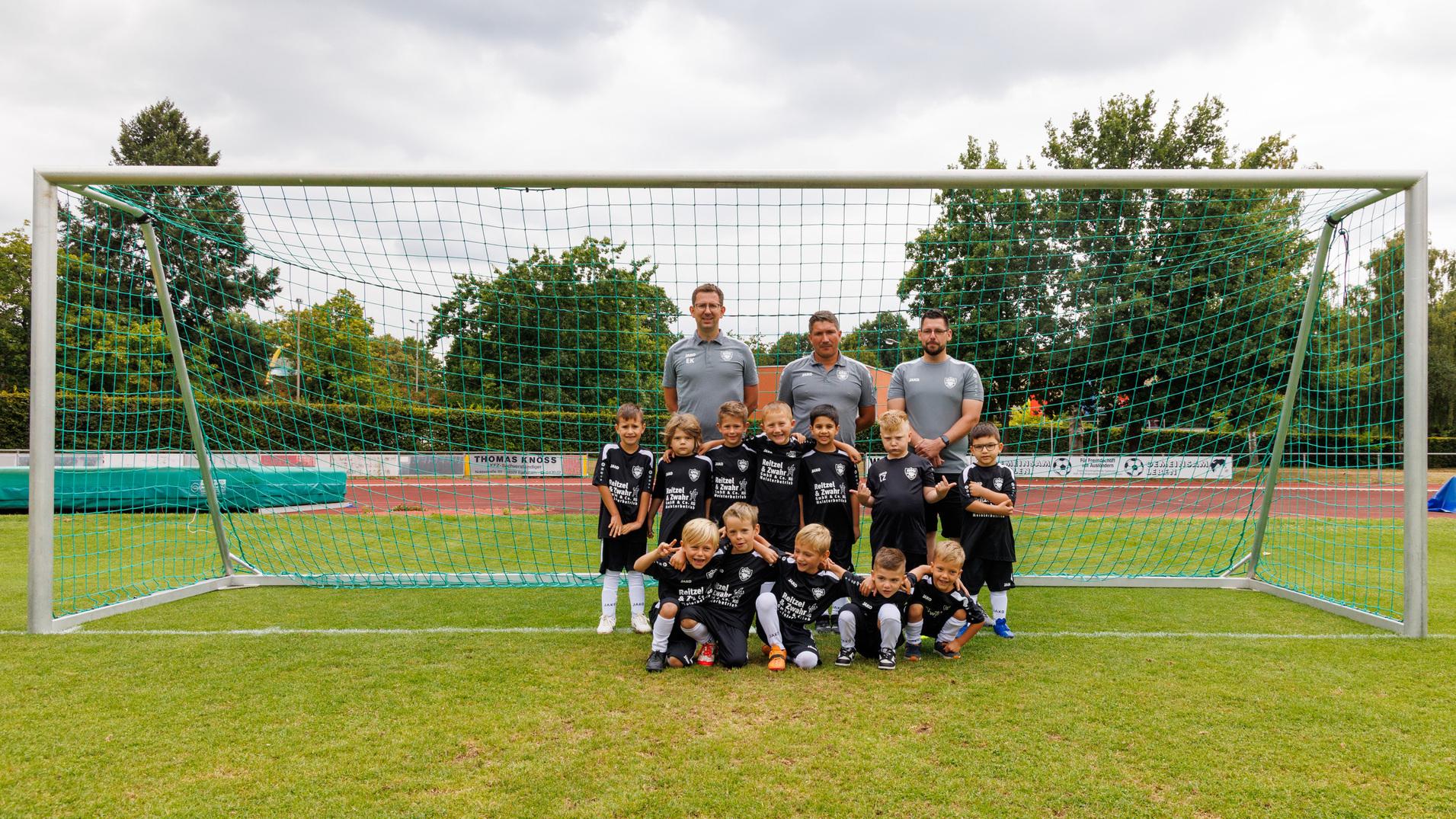 Kinderfußballmannschaft in schwarzen Trikots posiert vor einem Fußballtor auf einem Sportplatz. Erwachsene Trainer im Hintergrund.