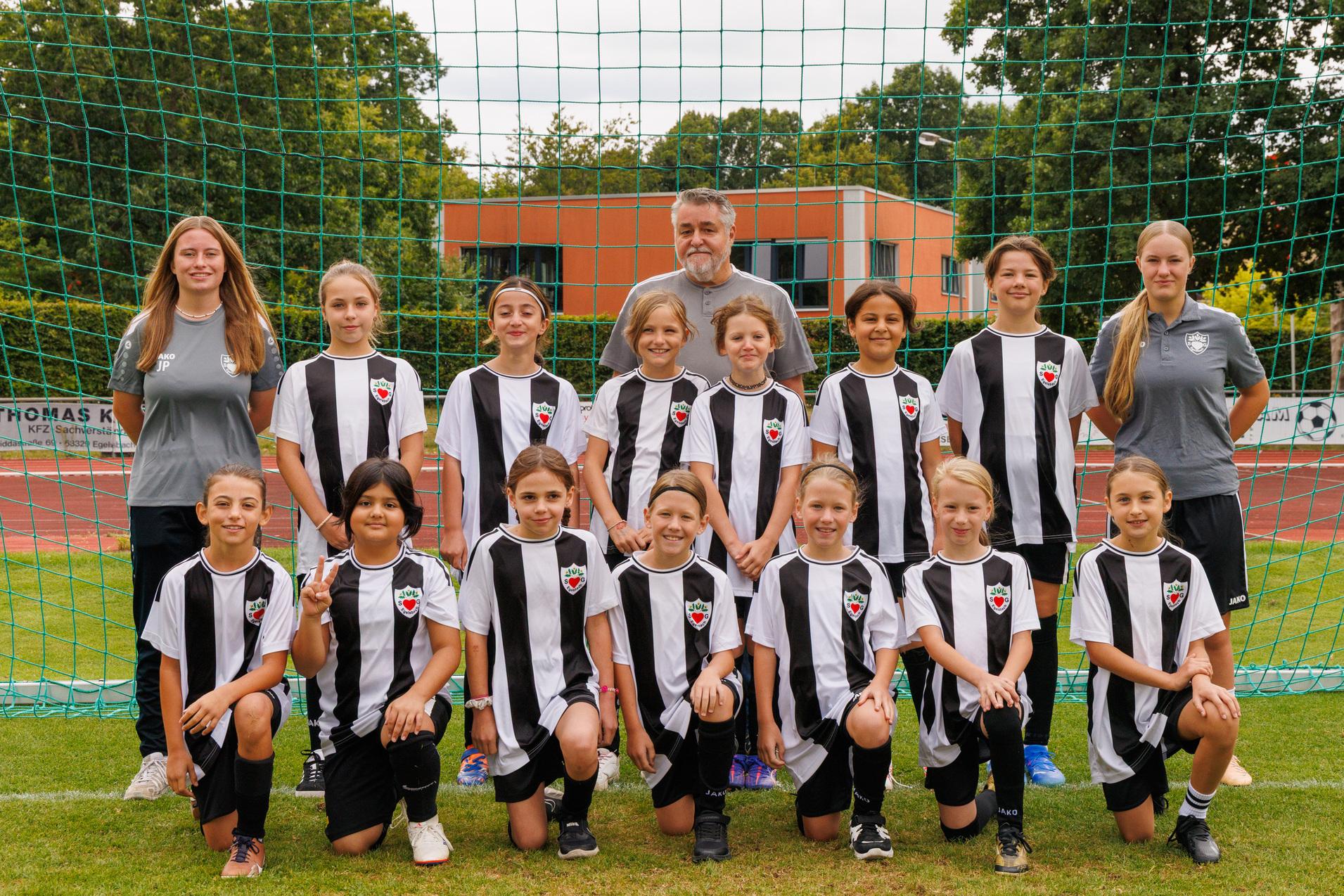 Gruppennahaufnahme eines Fu&szlig;ballteams mit M&auml;dchen in schwarz-wei&szlig;en Trikots und einem Trainer vor einem Fu&szlig;balltor.