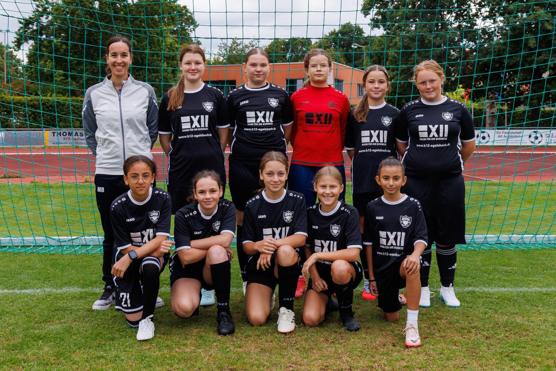 Mannschaftsfoto einer Fu&szlig;balljugend mit Trainerin, alle in schwarzen Trikots vor einem Fu&szlig;balltor.