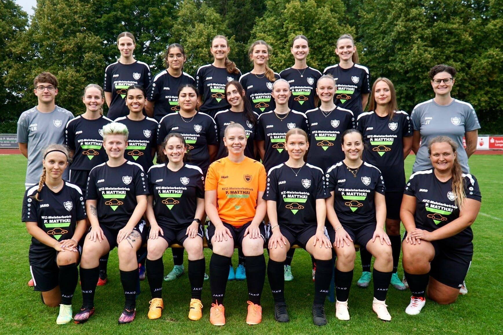 Gruppenfoto einer Frauenfußballmannschaft in schwarzen Trikots auf dem grünen Rasenplatz, 24 Spielerinnen und Trainer.