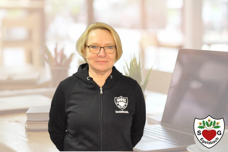 Frau mit blonden Haaren in schwarzer Jacke vor einem Laptop und Pflanzen im Hintergrund.