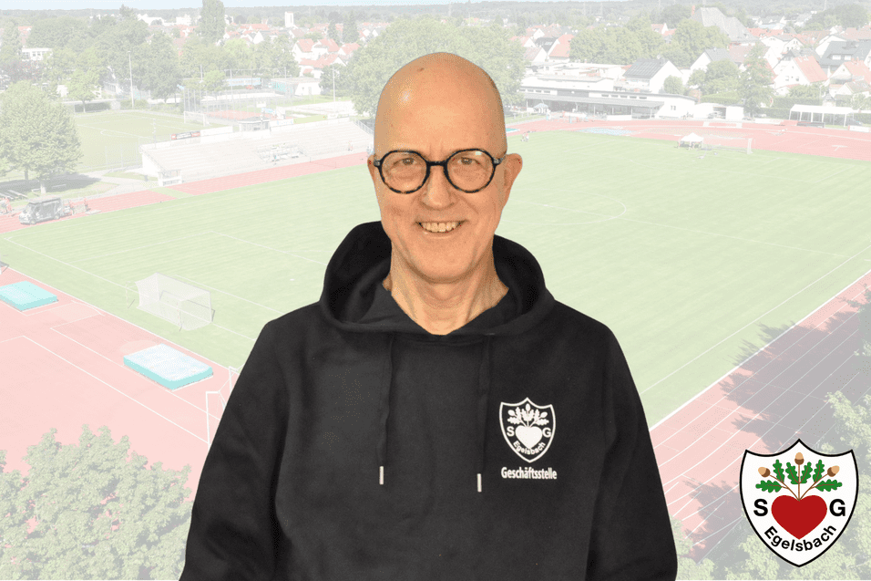 Mann mit Brille in Kapuzenpulli l&auml;chelt vor einem Sportplatz mit Rasen und Laufbahn. Logo unten rechts.