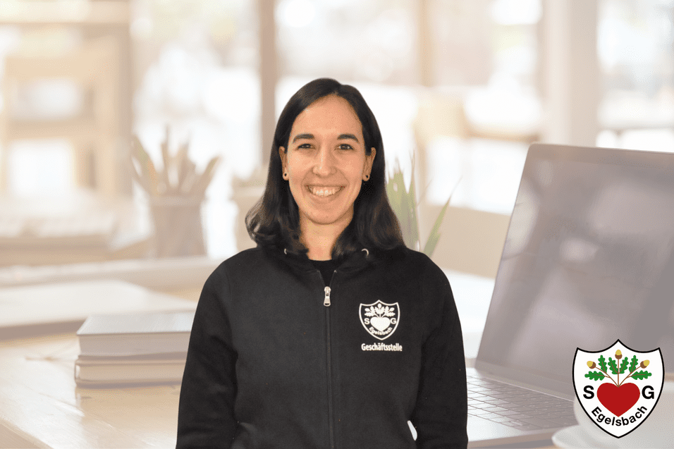 Frau mit schwarzer Sweatshirtjacke steht vor einem Schreibtisch mit Laptop und Pflanzen im Hintergrund.
