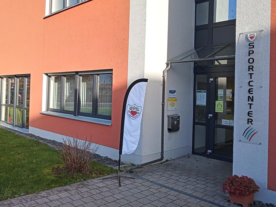 Eingang des Sportcenters mit gro&szlig;en Fenstern und einem Fahnenmast neben der T&uuml;r, umgeben von Rasen und Blumen.