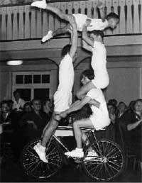 Drei Athleten f&uuml;hren eine akrobatische Darbietung auf einem Fahrrad in einem festlichen Raum aus.