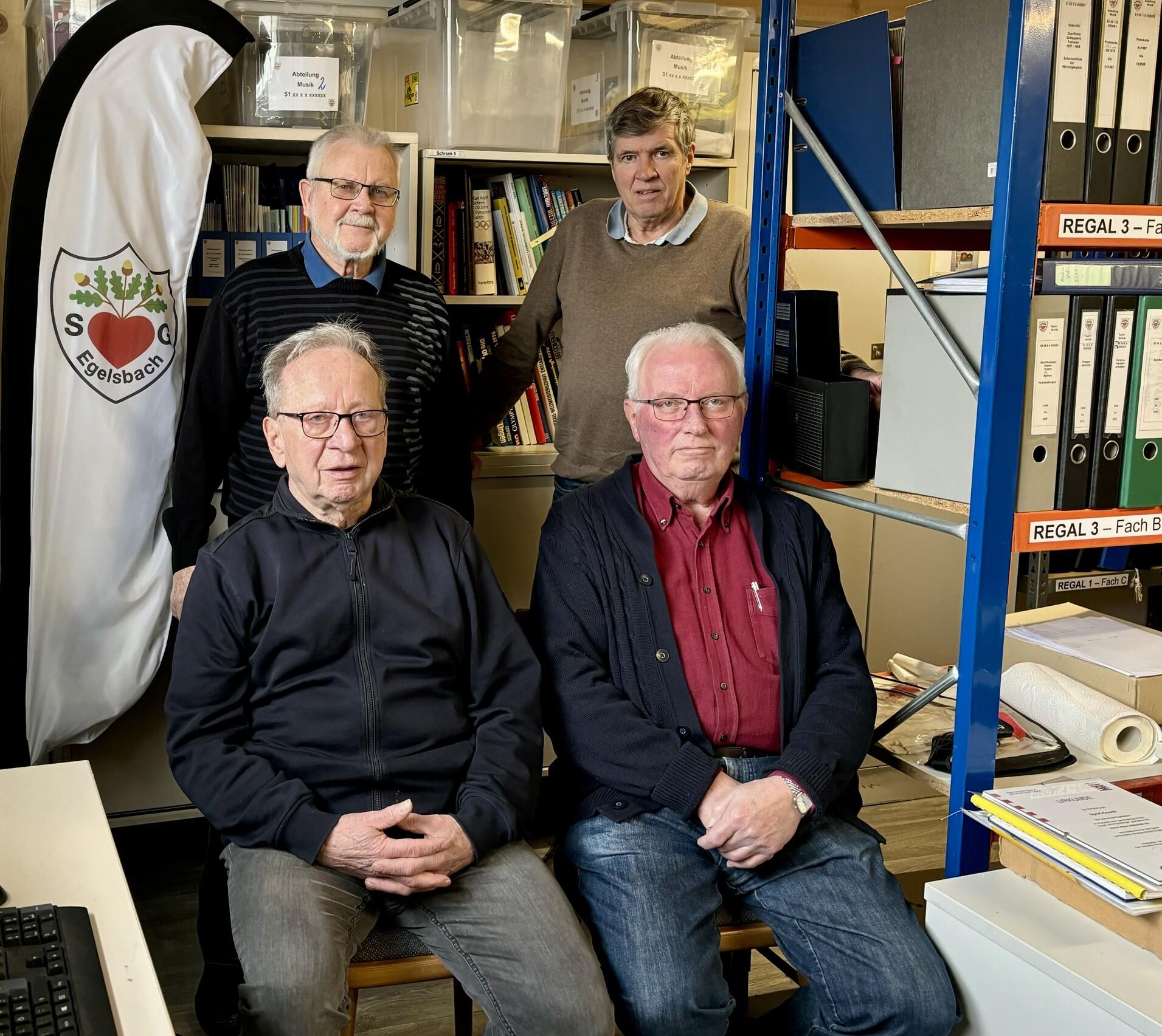 Vier Männer sitzen und stehen in einem Raum mit Regalen, Bücher und einem Vereinswimpel im Hintergrund.
