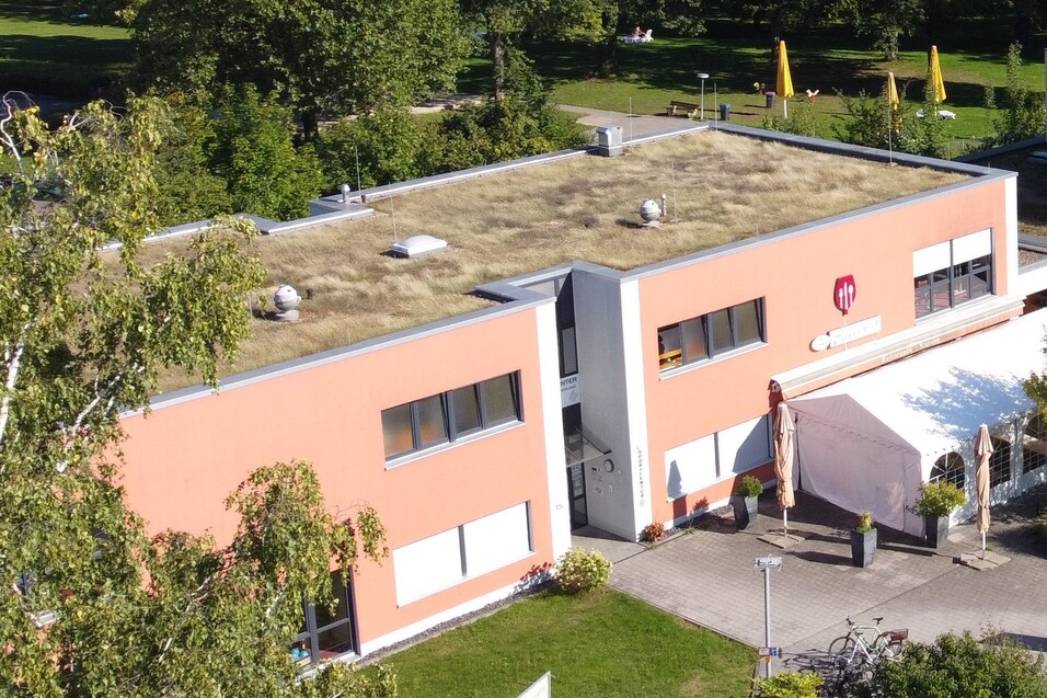 Dachansicht eines flachgedeckten Geb&auml;udes mit begr&uuml;ntem Dach und hellorangefarbenen W&auml;nden, umgeben von B&auml;umen.